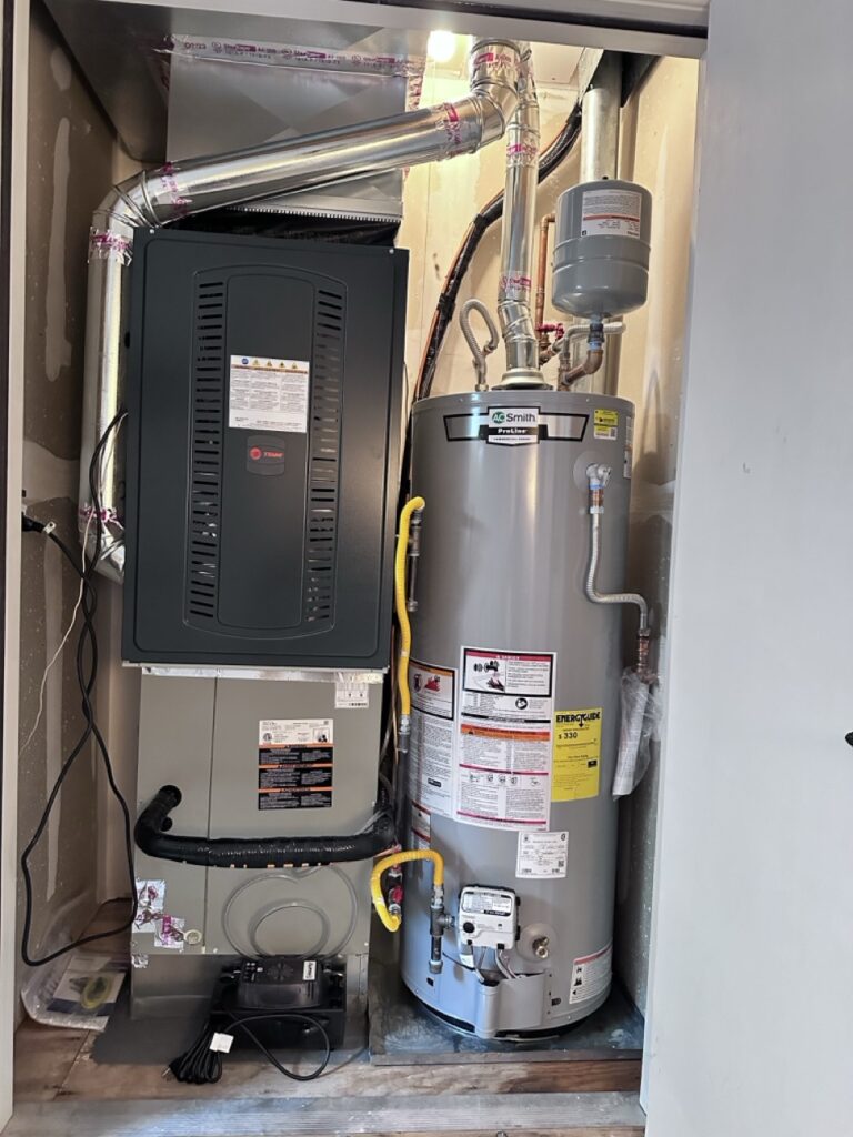 A utility closet containing a black Trane air handler/furnace on the left and a gray A.O. Smith ProLine gas water heater on the right, with metal venting, copper piping, and a condensate pump.