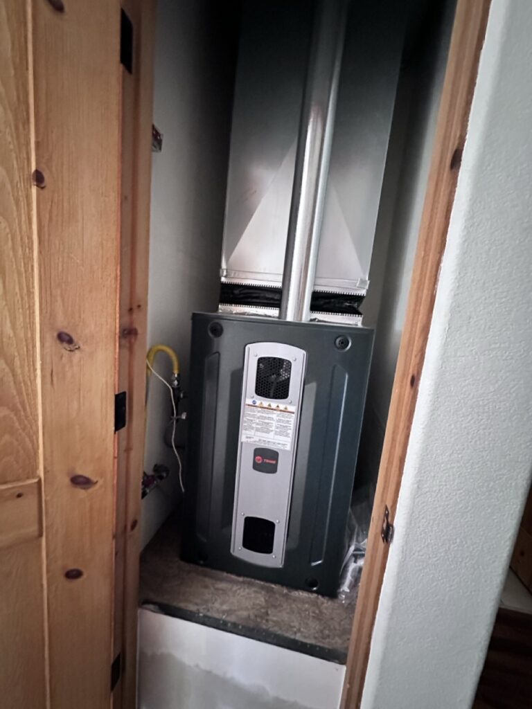 A Trane gas furnace installed in a narrow closet with wood door frame, ductwork above, and a yellow gas line connection on the left.